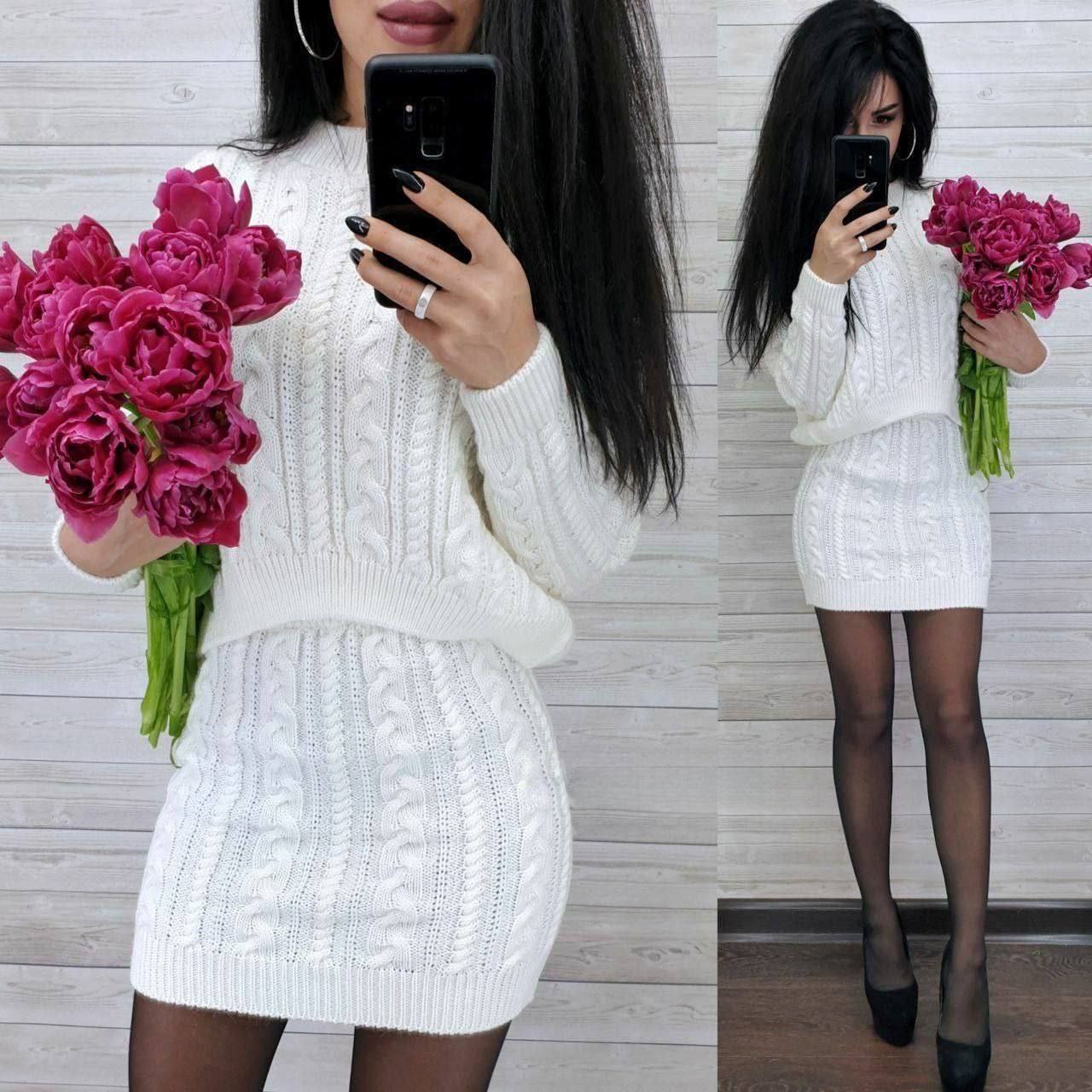 Woman in a white knitted outfit holding pink flowers, taking a mirror selfie.