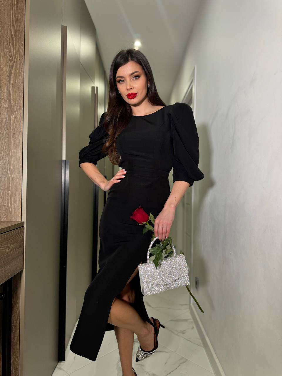 Woman in a black dress holding a red rose and a silver handbag in a hallway.