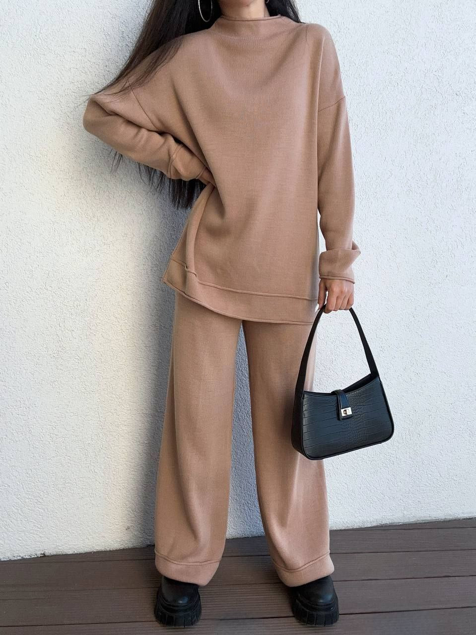 Person wearing a beige outfit holding a black handbag against a white wall.