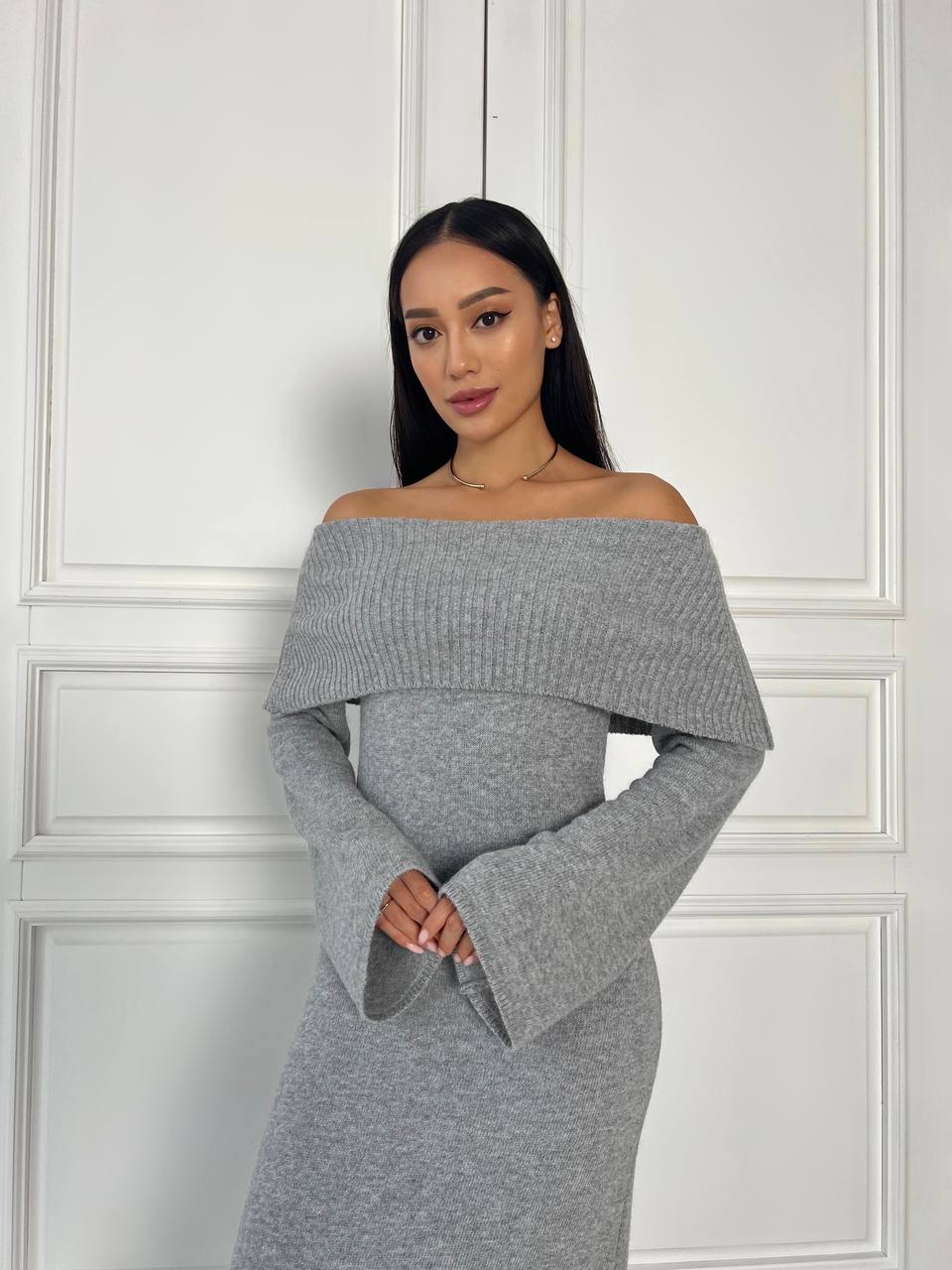 Woman wearing a gray off-shoulder dress against a white paneled wall.