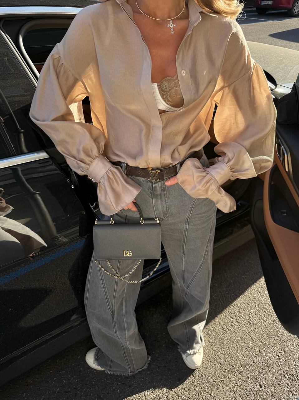 Person wearing a beige blouse and jeans with a black handbag, standing next to a car.