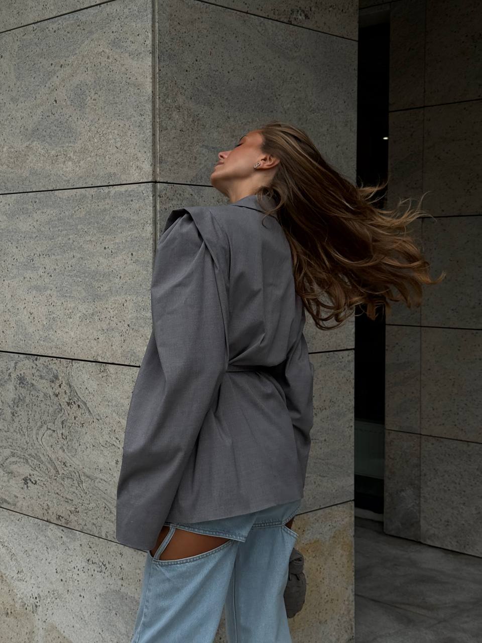 Person wearing a gray coat and light blue jeans against a stone wall.