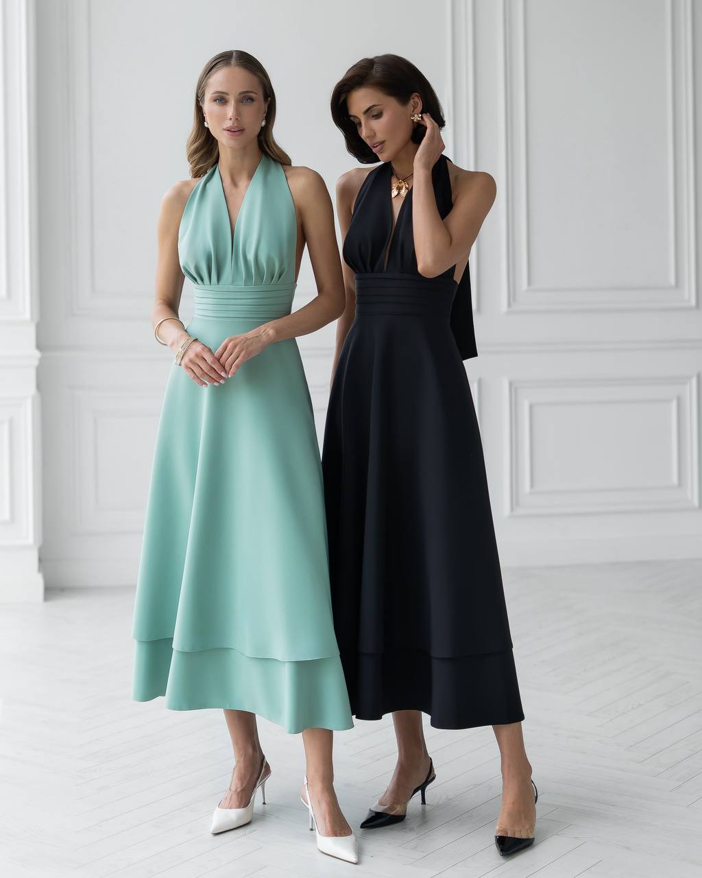 Two women wearing elegant dresses standing against a white wall.