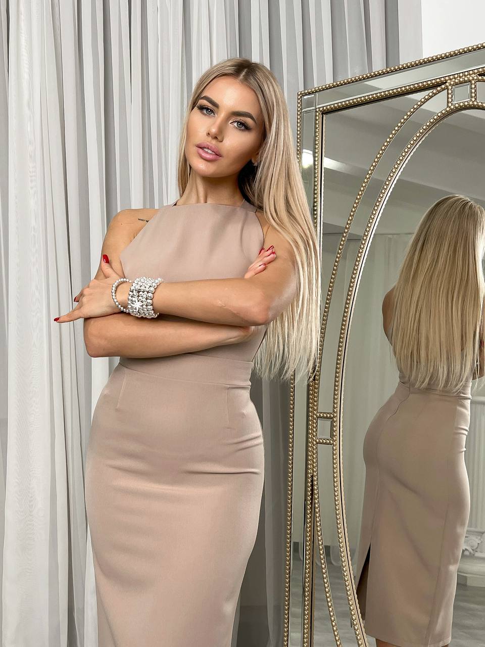 Woman in a beige dress standing in front of a mirror with white curtains.