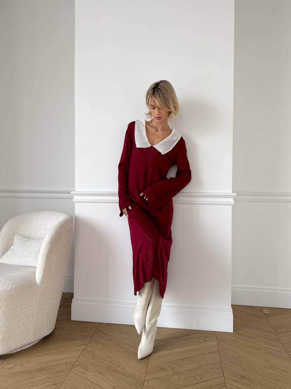 Woman in a burgundy dress with white collar standing against a white wall.