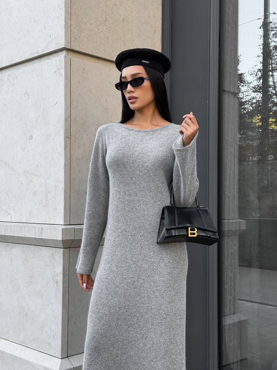 Woman in a gray dress and black hat holding a black handbag against a building.