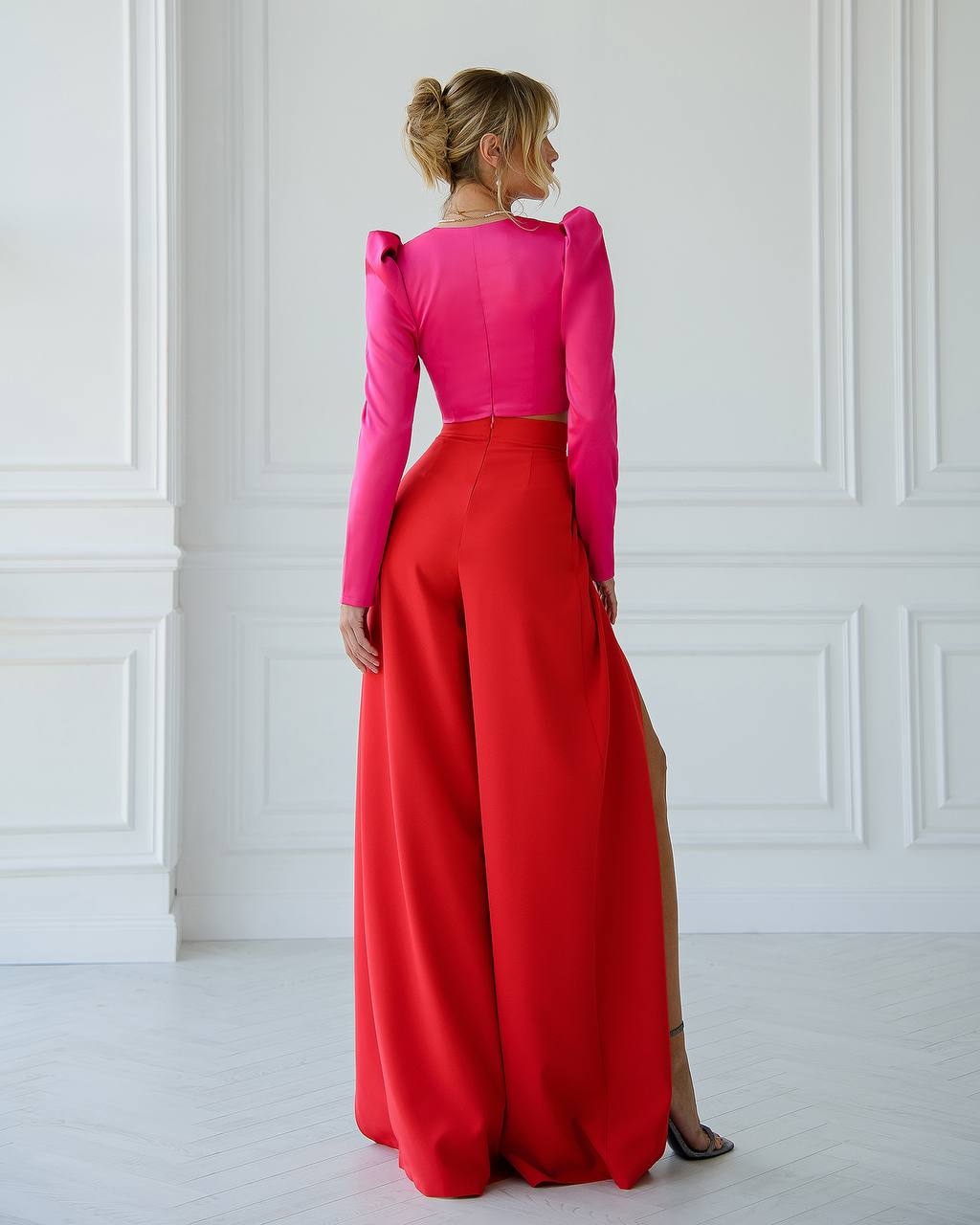 Woman wearing a bright pink top and red skirt against a white wall.
