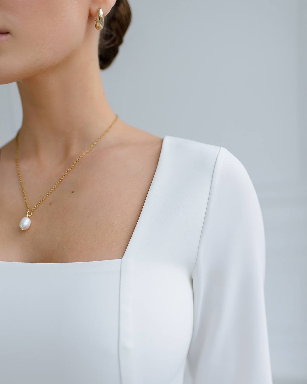 Person wearing a gold necklace with a pearl pendant against a neutral background