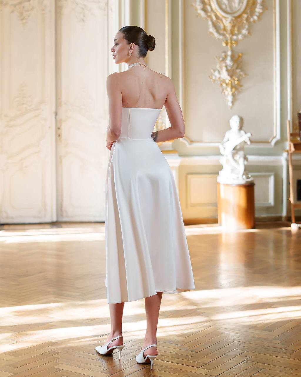 Woman in a white dress standing in an elegant room with classical decor.