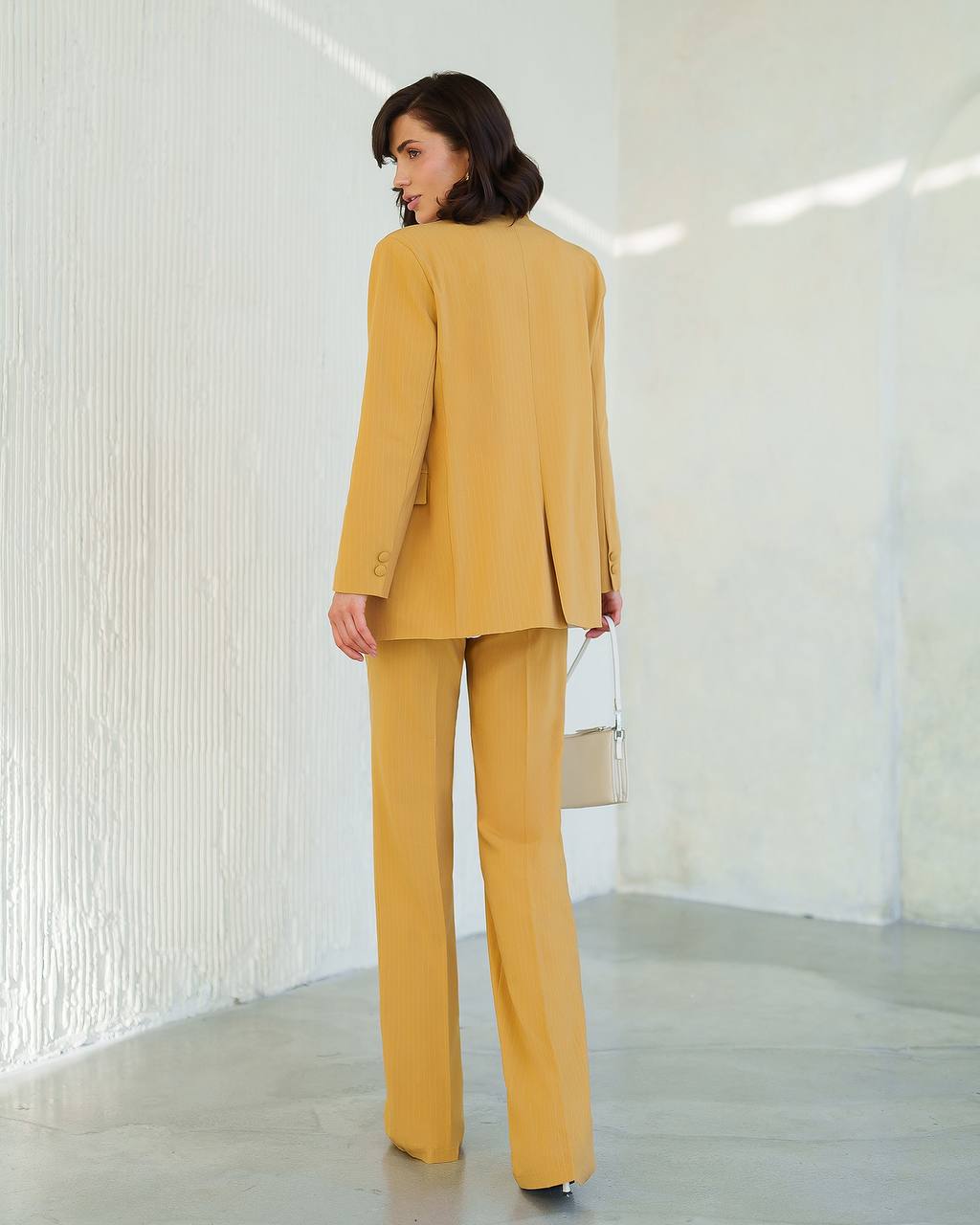 Woman wearing a mustard yellow suit standing in a minimalistic room with white walls.