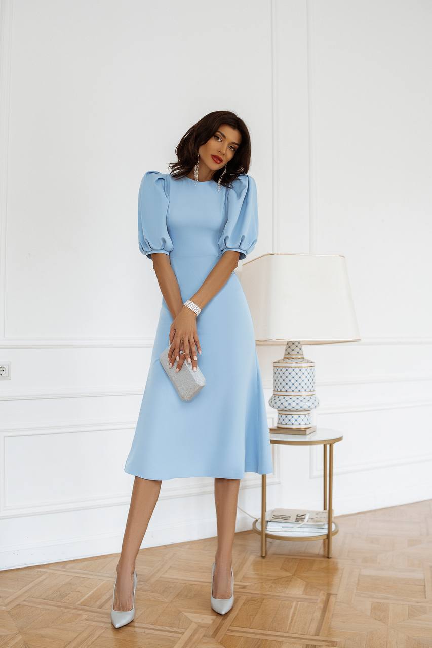 Woman in a light blue dress standing in a room with a white wall and wooden floor.