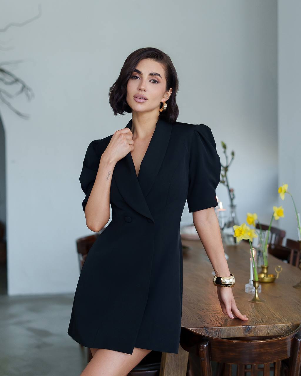 Woman in a black dress standing in a room with a wooden table and flowers.