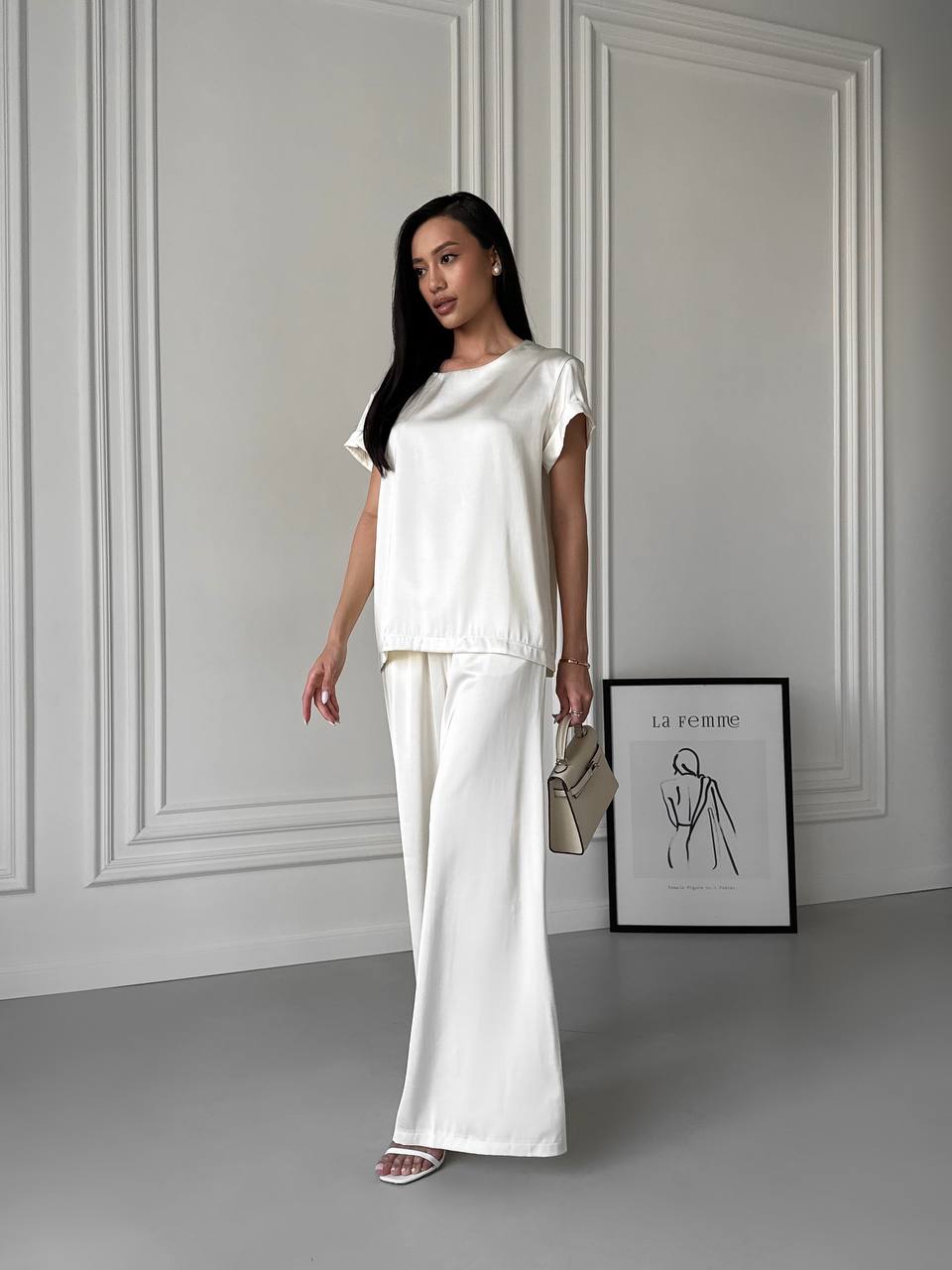 Woman in a white outfit standing in a minimalistic room with a framed picture on the wall.