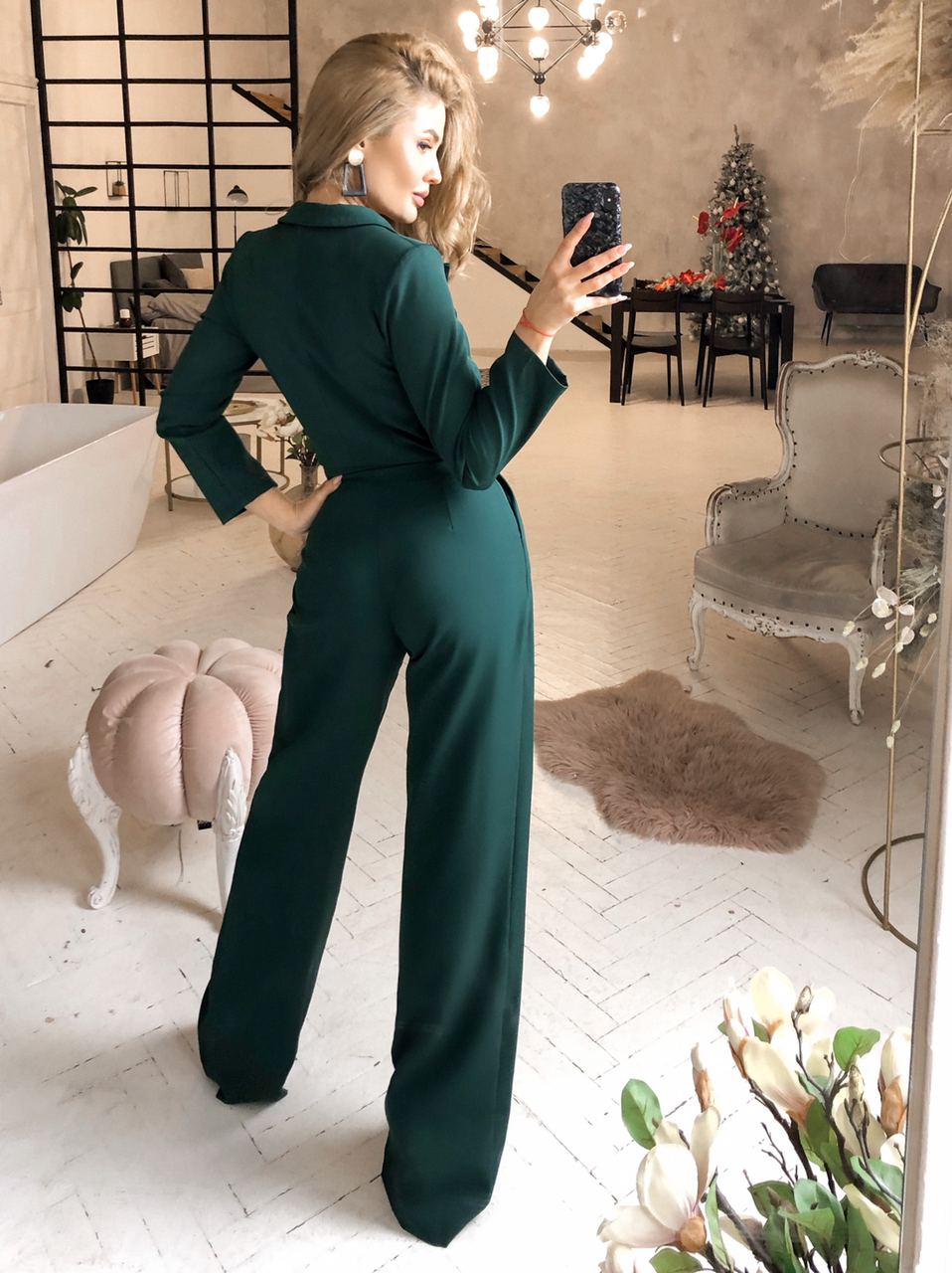 Woman in a green jumpsuit taking a mirror selfie in a stylish room.