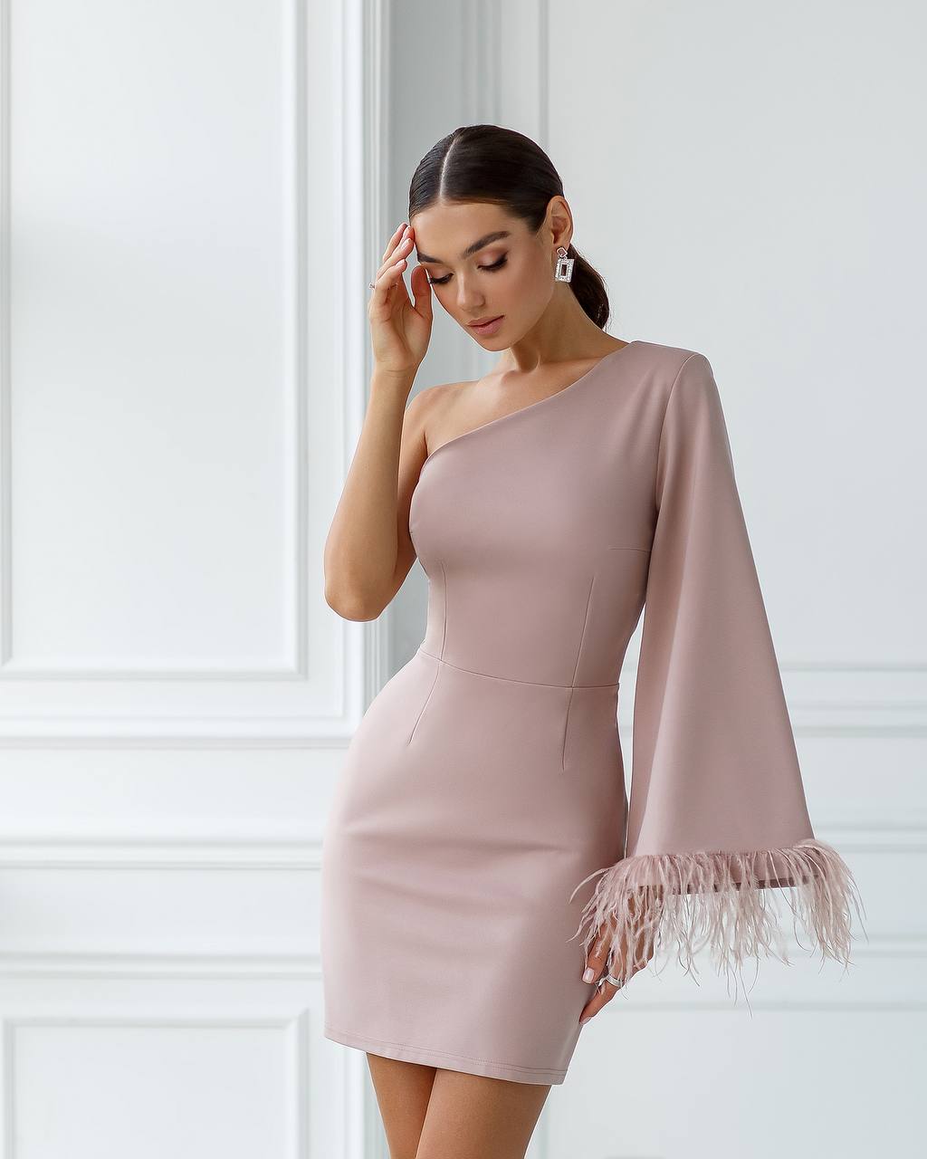Woman wearing a one-shoulder pink dress with fringe details against a white wall.