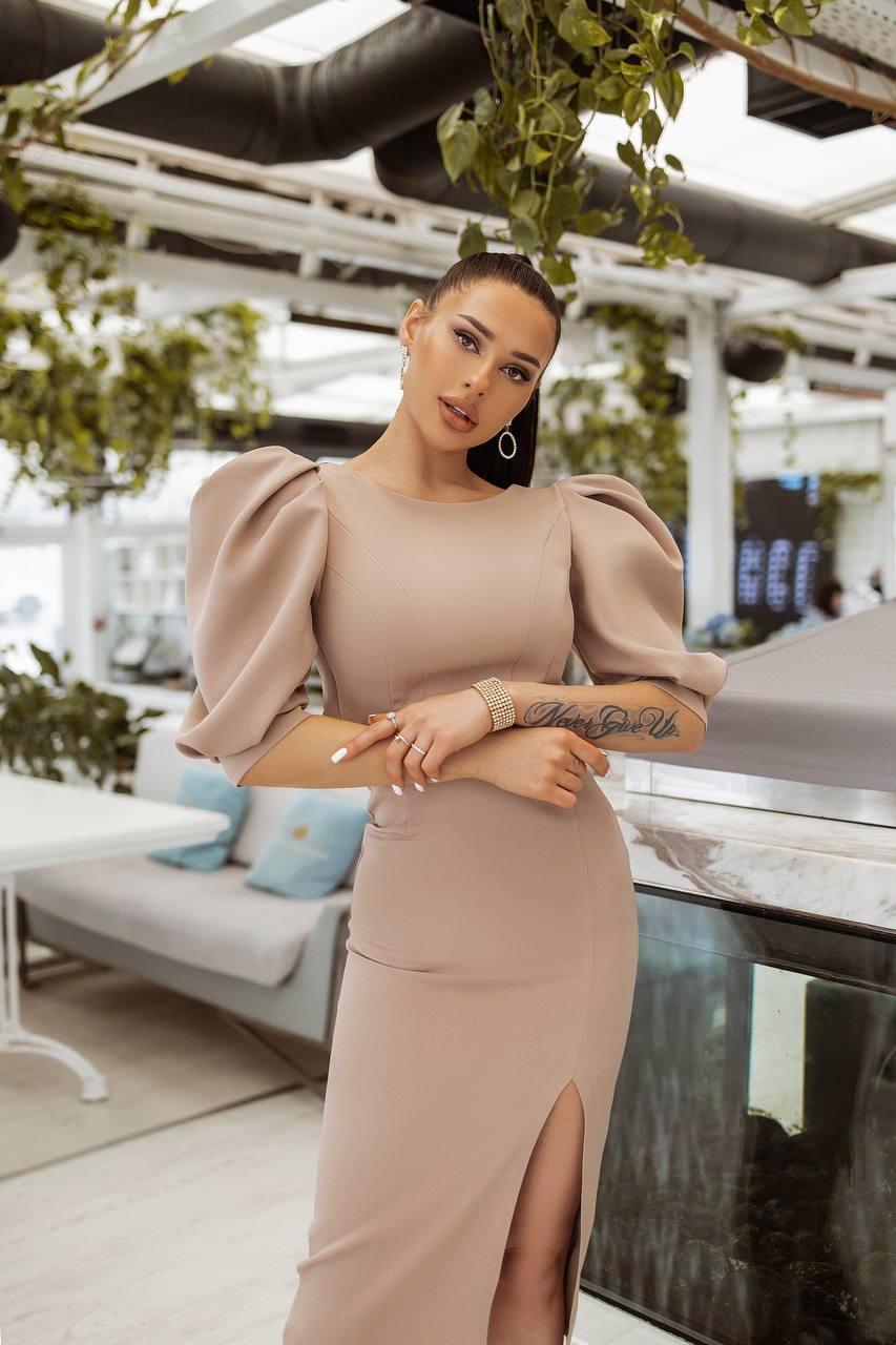 Woman in a beige dress standing in an outdoor setting with plants and furniture.