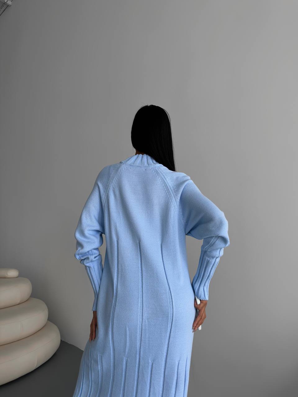 Person wearing a light blue dress standing against a plain wall with a beige sculpture in the corner.