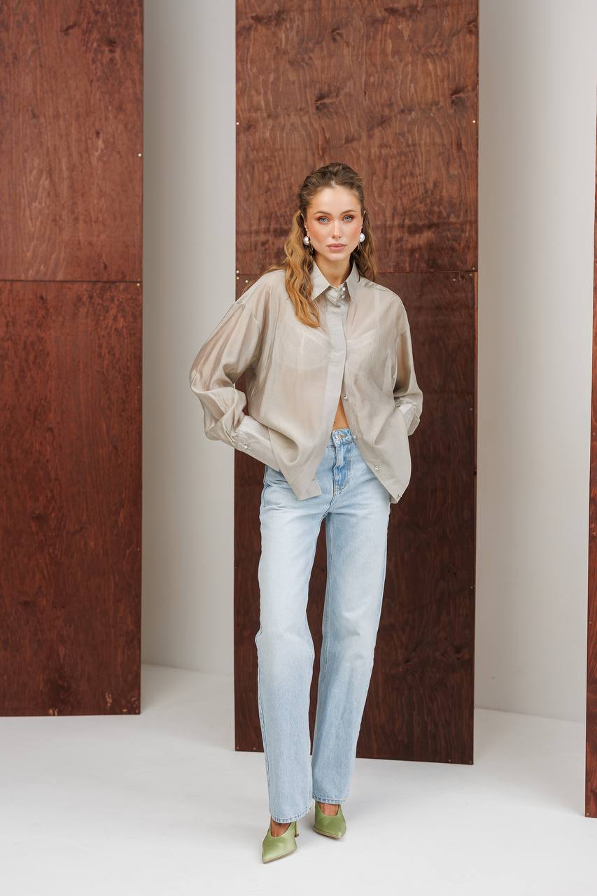 Woman wearing a beige blouse and light blue jeans standing against a wooden panel background