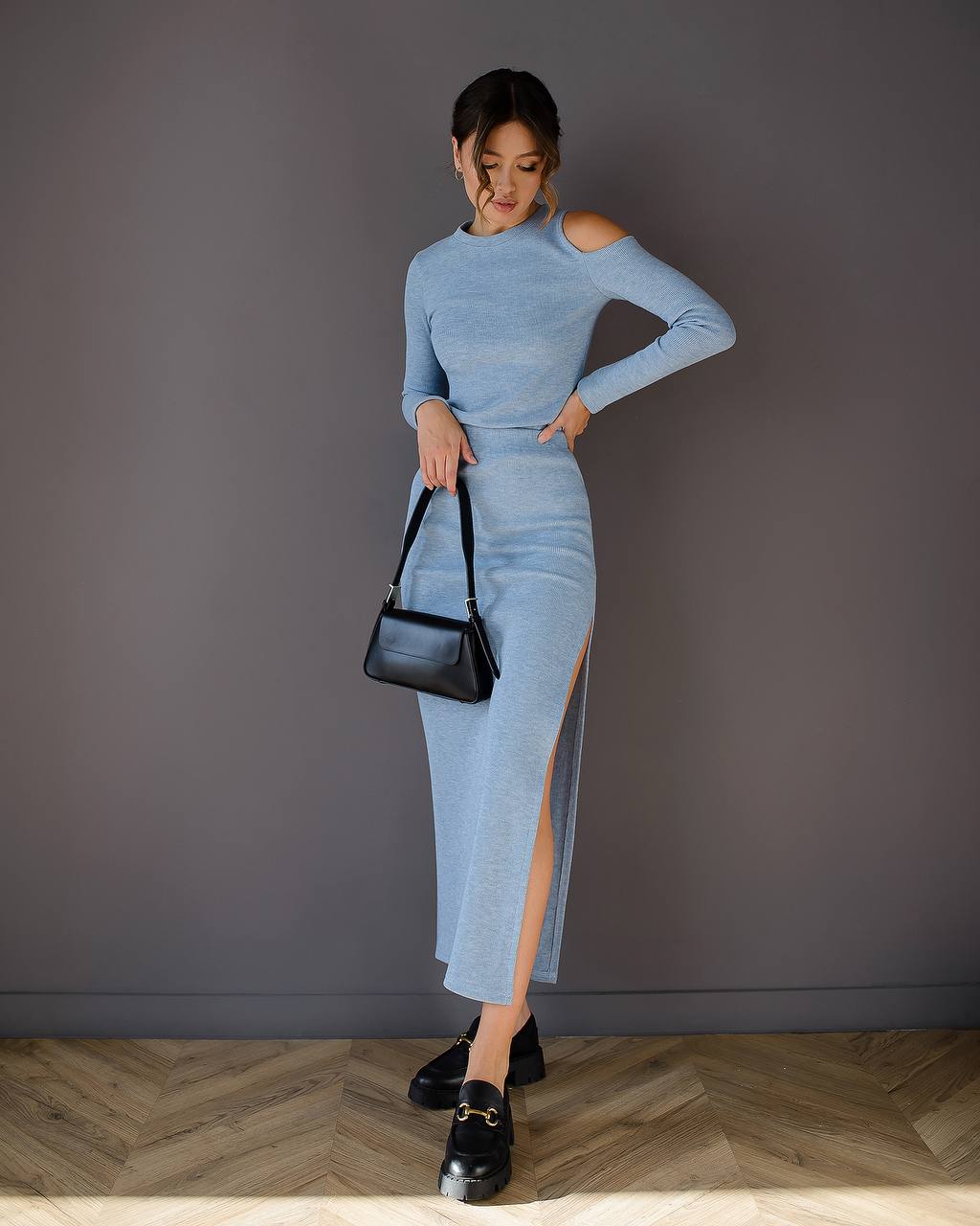 Woman wearing a blue dress with a black handbag against a gray wall.