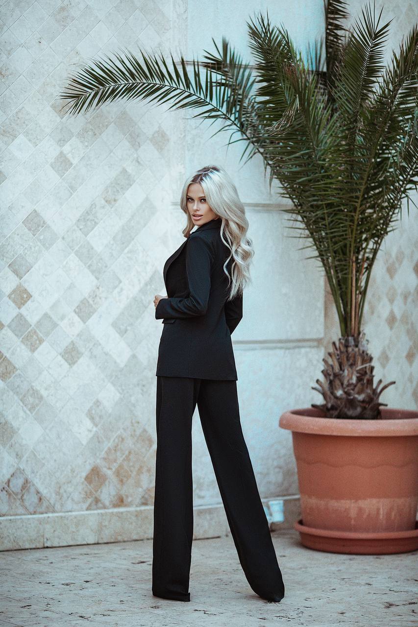 Woman in a black suit standing next to a potted plant against a textured wall.