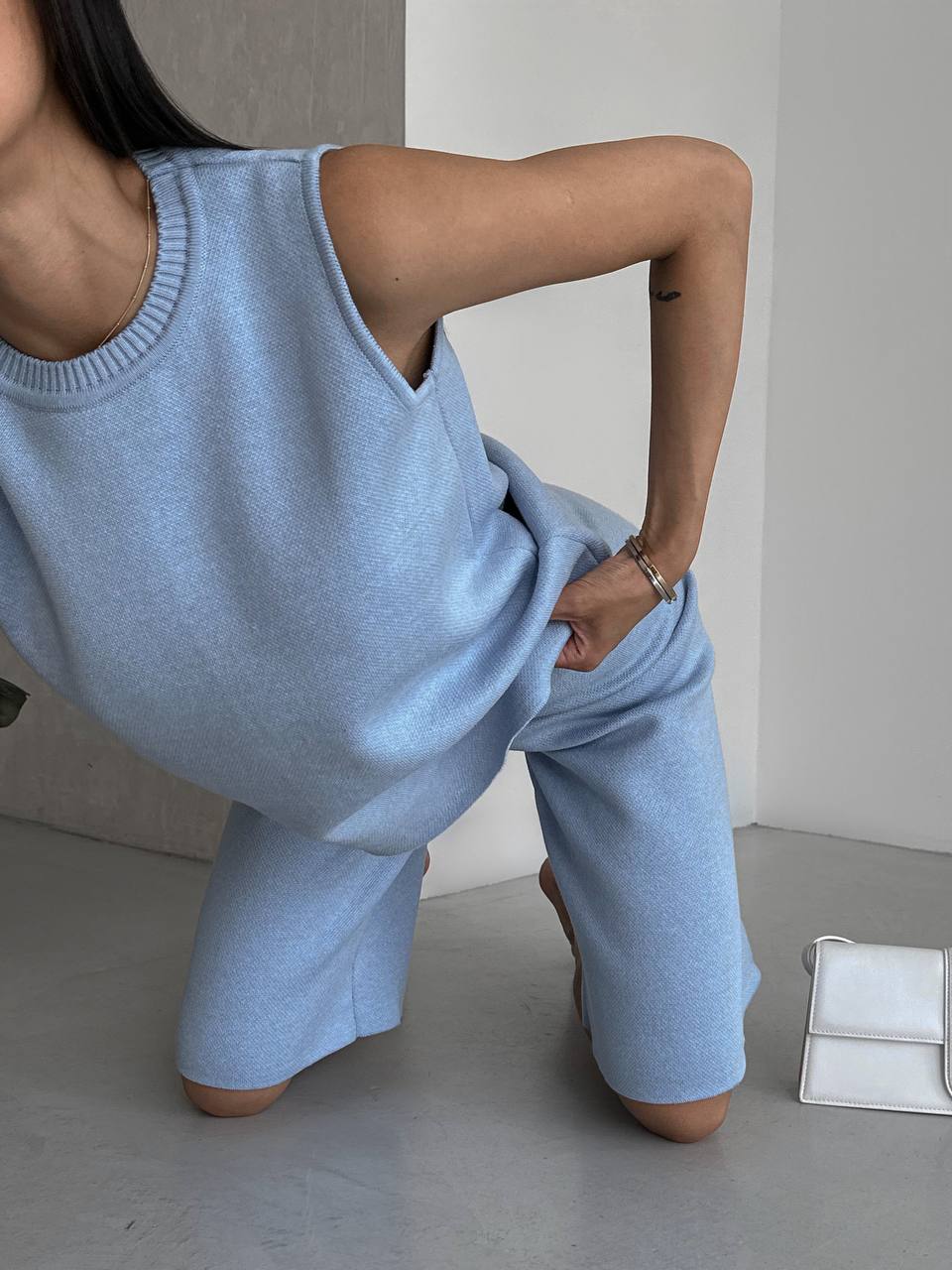 Person wearing a light blue sleeveless top and pants in a minimalistic setting.