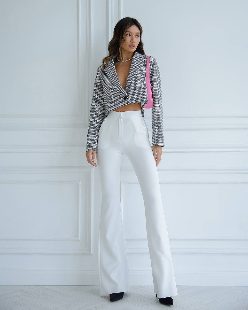 Woman wearing a checkered blazer and white pants with a pink bag against a white wall.