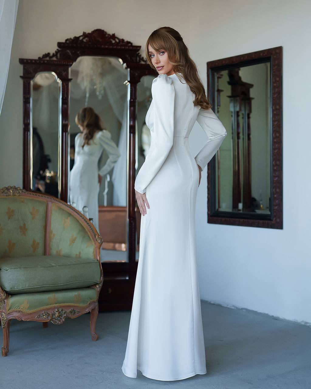 Woman in a white dress standing in a room with mirrors and a vintage chair.