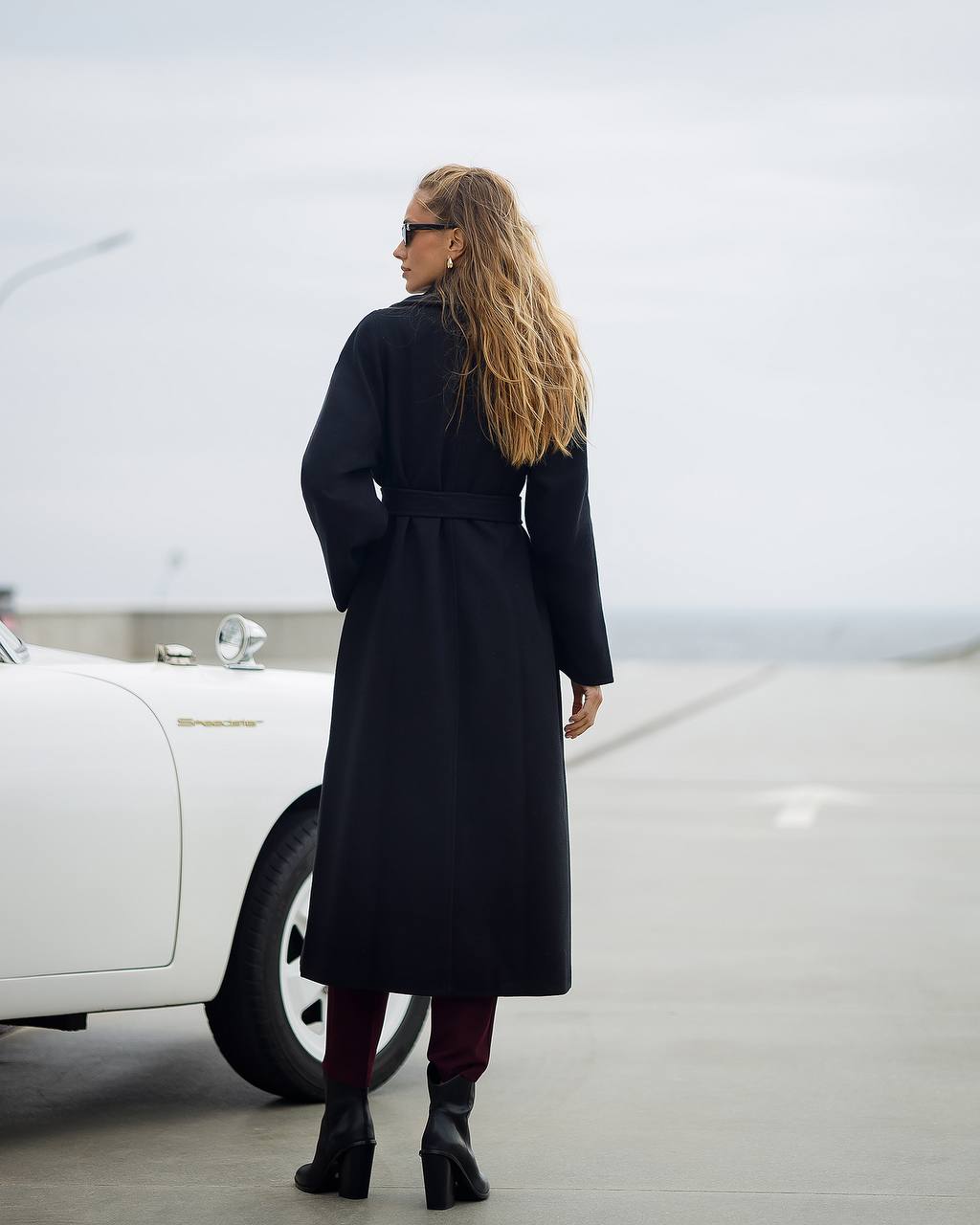 Woman in a long black coat standing next to a white car on a road.