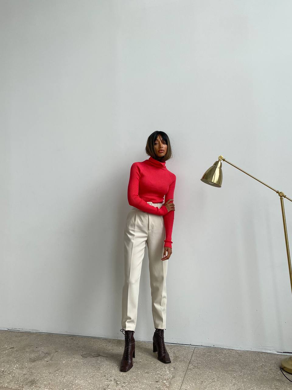 Person wearing a red turtleneck and white pants standing against a plain wall with a gold lamp.