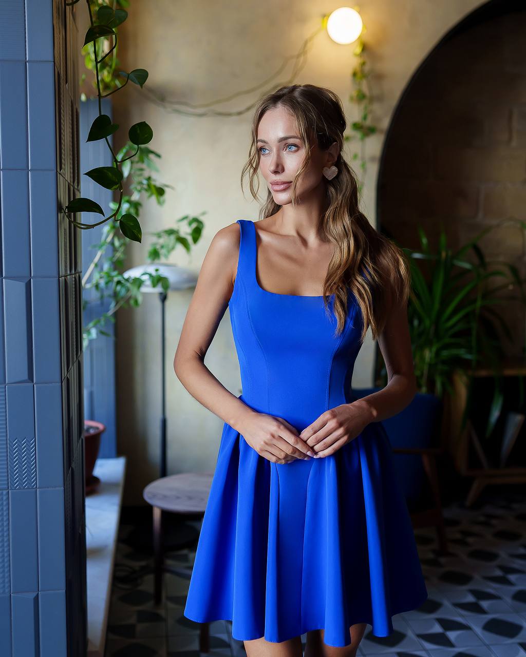 Woman in a blue dress standing in an indoor setting with plants and a blue wall.