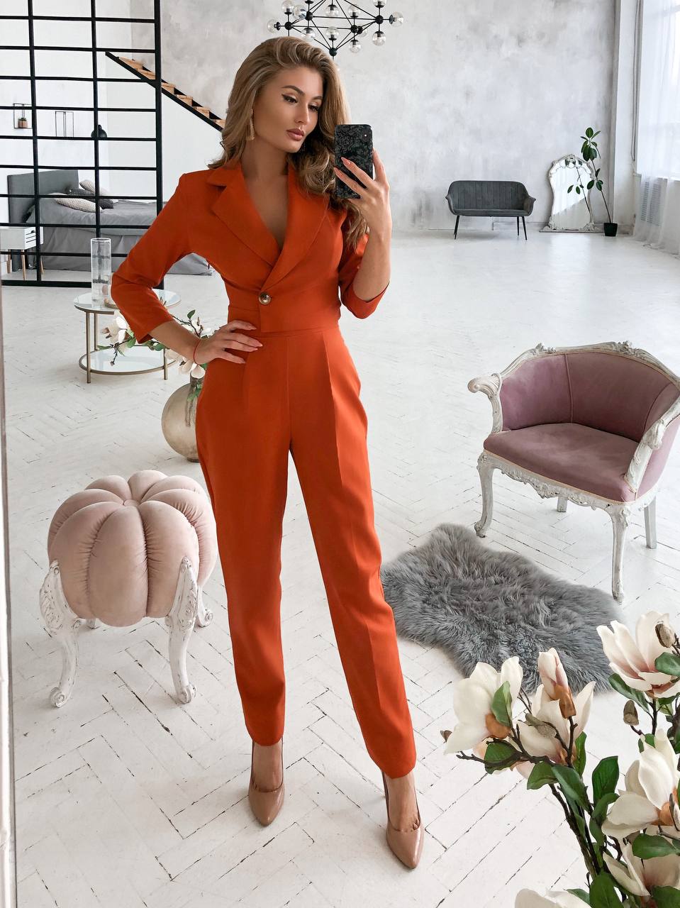 Woman in an orange jumpsuit taking a mirror selfie in a stylish room.