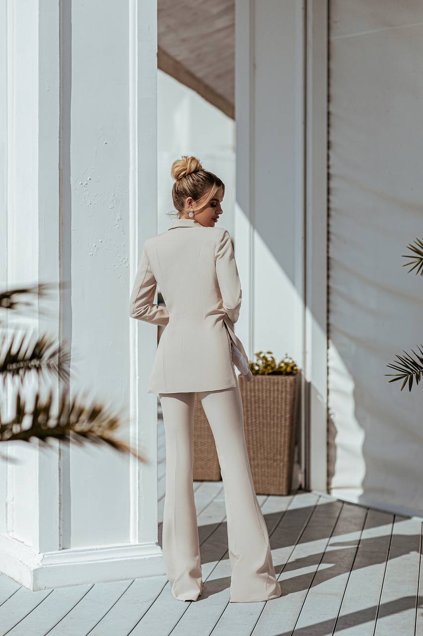 Woman in a beige suit standing on a wooden deck with white walls and plants.