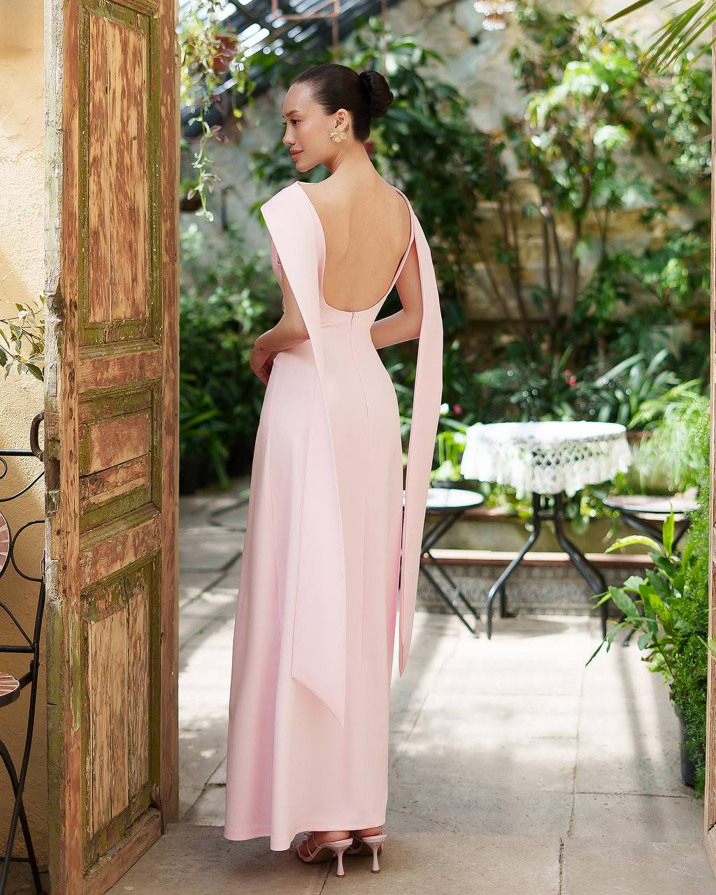 Woman in a pink gown with an open back standing in a garden setting.