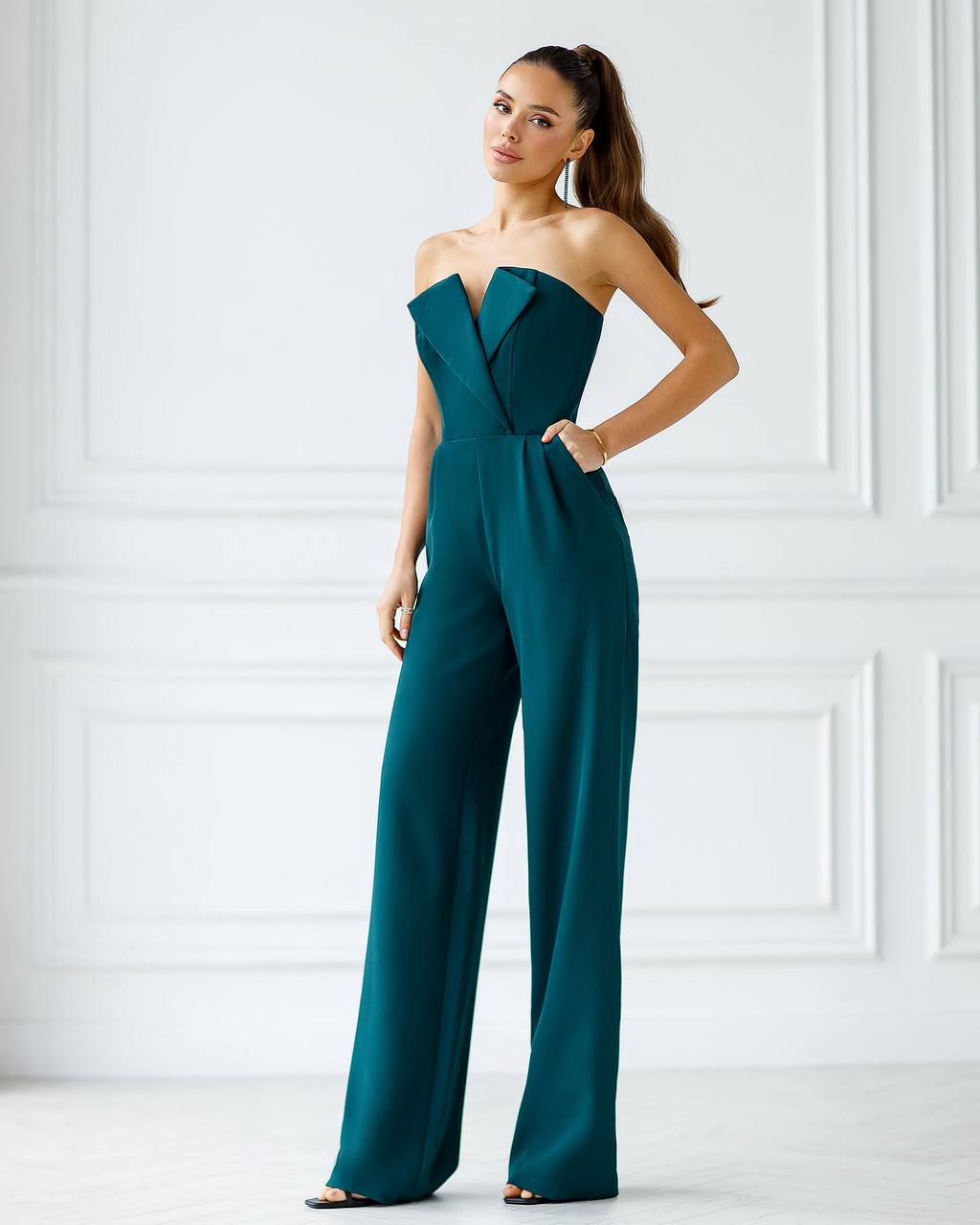 Woman wearing a teal strapless jumpsuit against a white wall