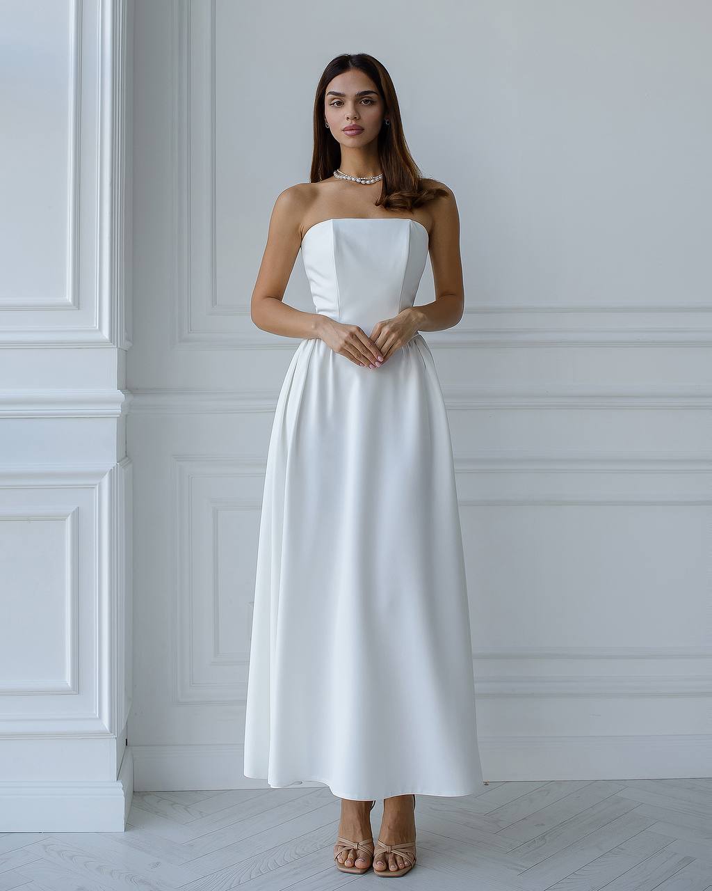 Woman wearing a white strapless dress standing against a white wall.