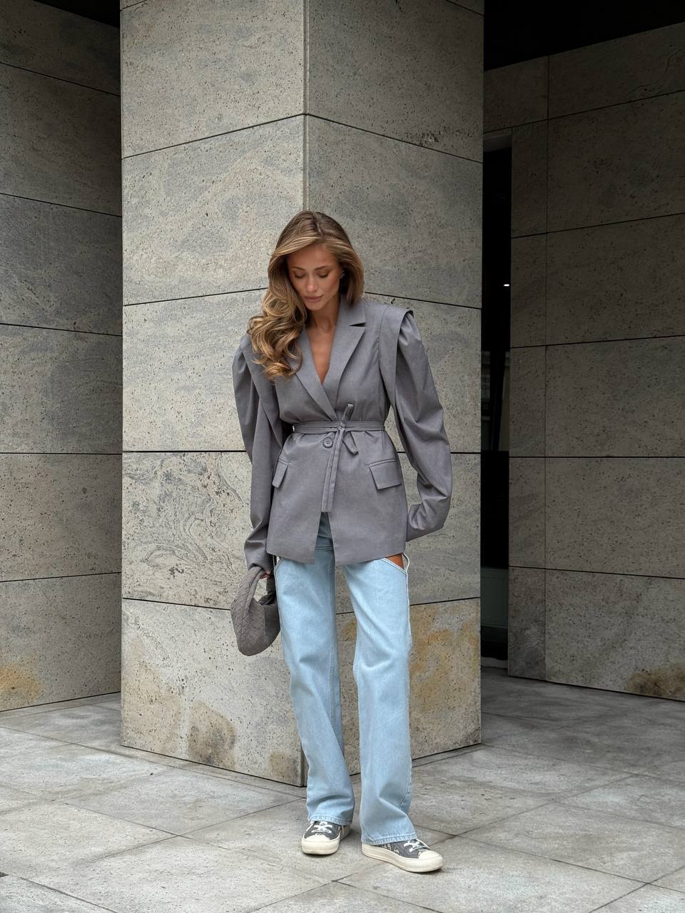 Woman wearing a gray blazer and light blue jeans standing against a stone wall.