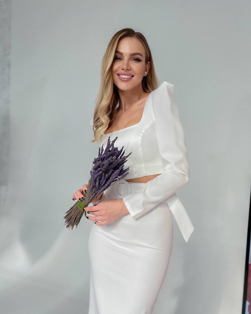 Woman in a white dress holding a bouquet of lavender against a plain background
