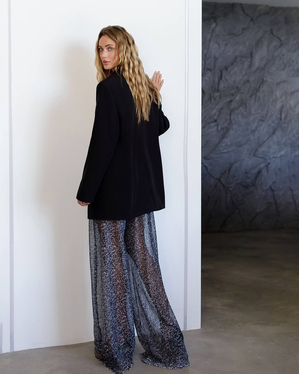 Woman wearing a black jacket and patterned pants standing against a white wall.
