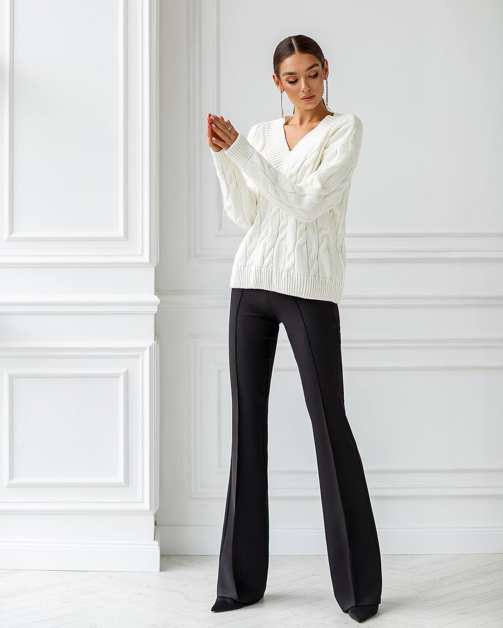 Woman wearing a white cable knit sweater and black pants against a white wall.