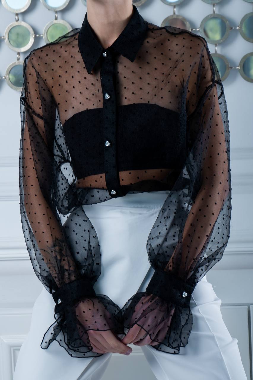 Person wearing a sheer black polka dot blouse over a white outfit with a decorative wall in the background.