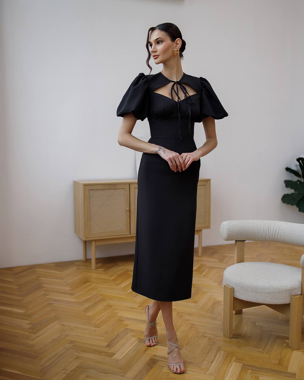 Woman in a black dress standing in a room with wooden flooring and a white wall.