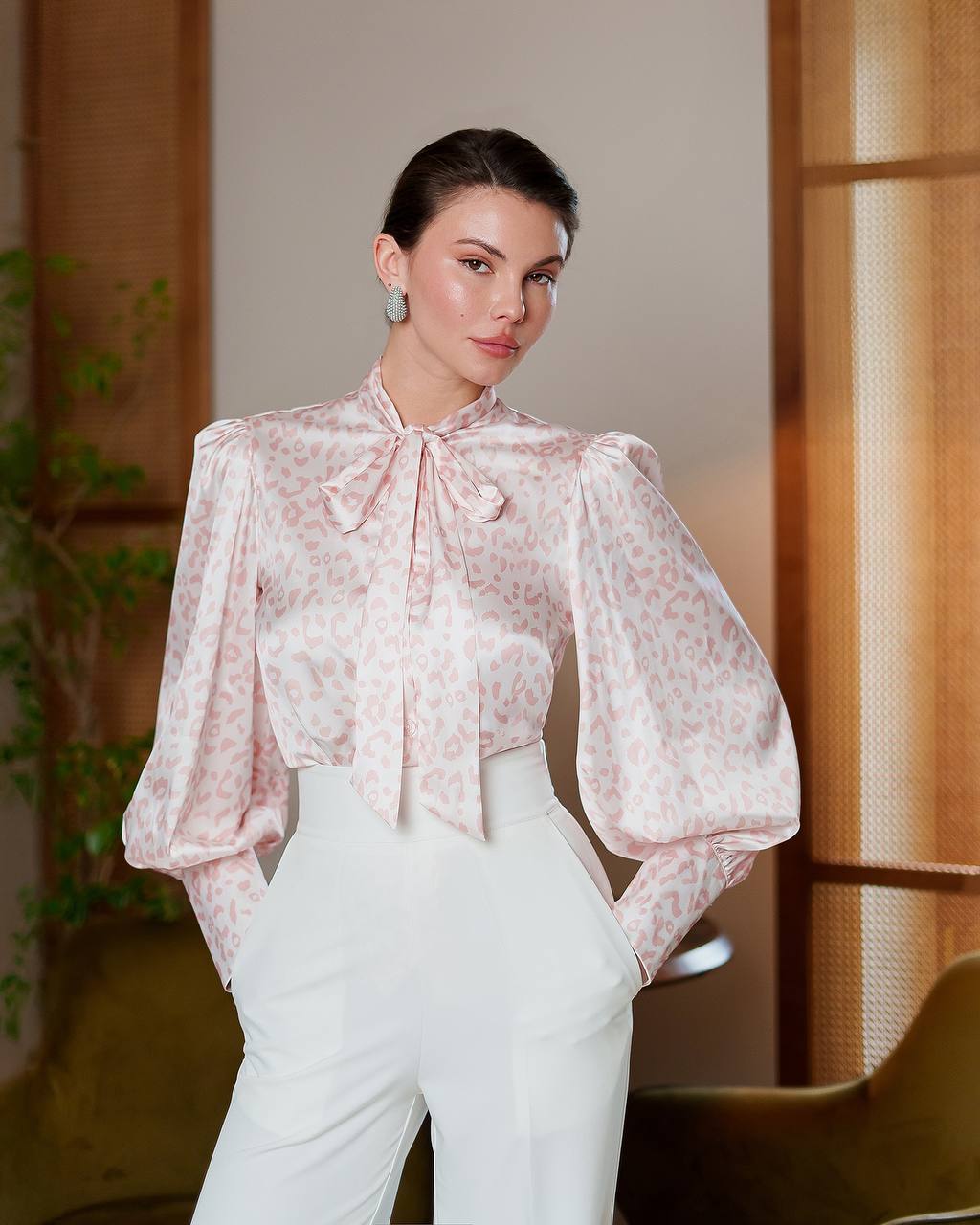 Woman wearing a light pink blouse with a bow tie and white pants in an indoor setting.