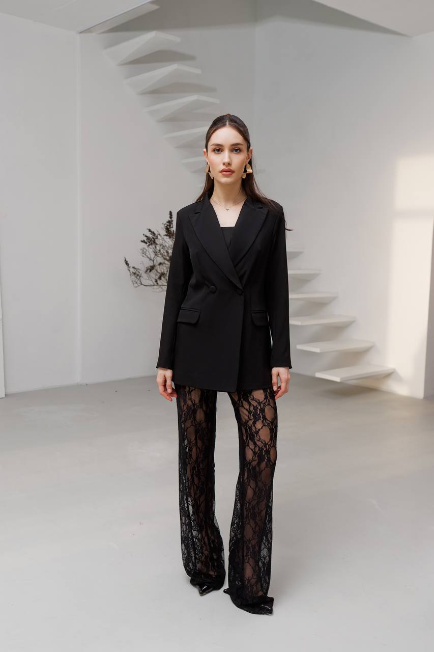 Woman wearing a black suit with sheer black pants in a minimal white room.