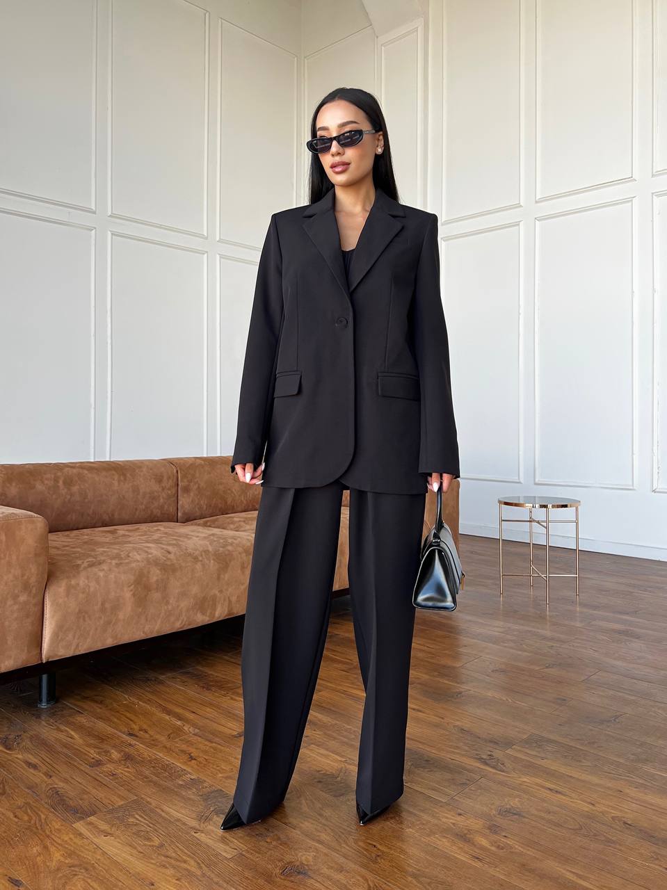 Woman in a black suit standing in a modern room with wooden flooring and a brown sofa.