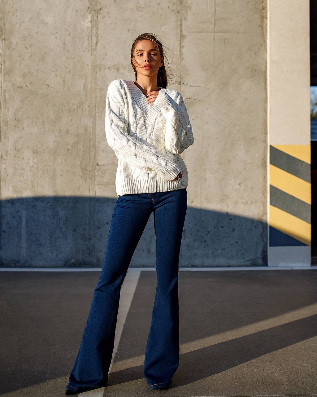 Woman wearing a white sweater and blue flared jeans standing against a concrete wall.