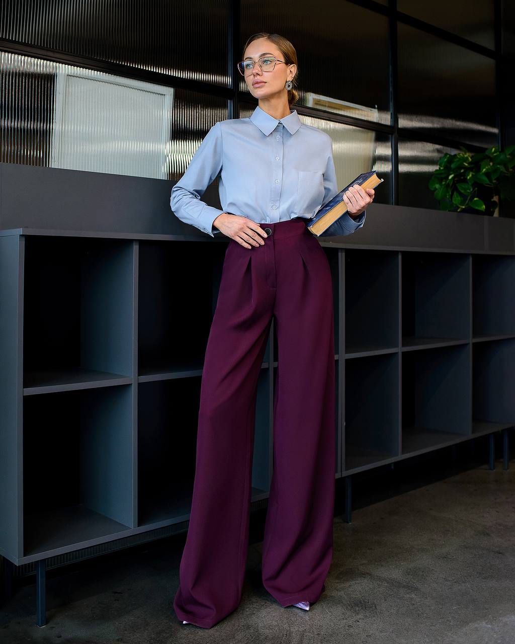 Woman in a light blue shirt and purple pants standing in an office setting.