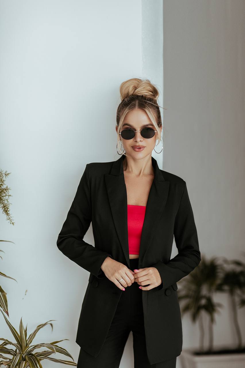 Woman in a black suit with a red top standing against a light-colored wall with plants.