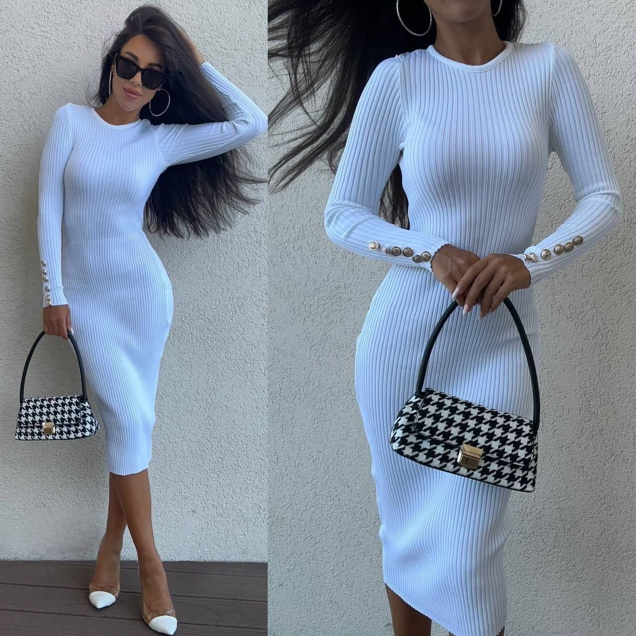 Woman wearing a white ribbed dress holding a black and white checkered handbag against a plain background.