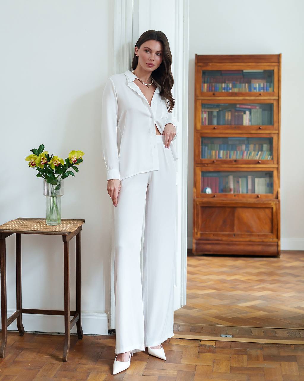 Woman in a white outfit standing in a room with wooden flooring and a bookshelf.