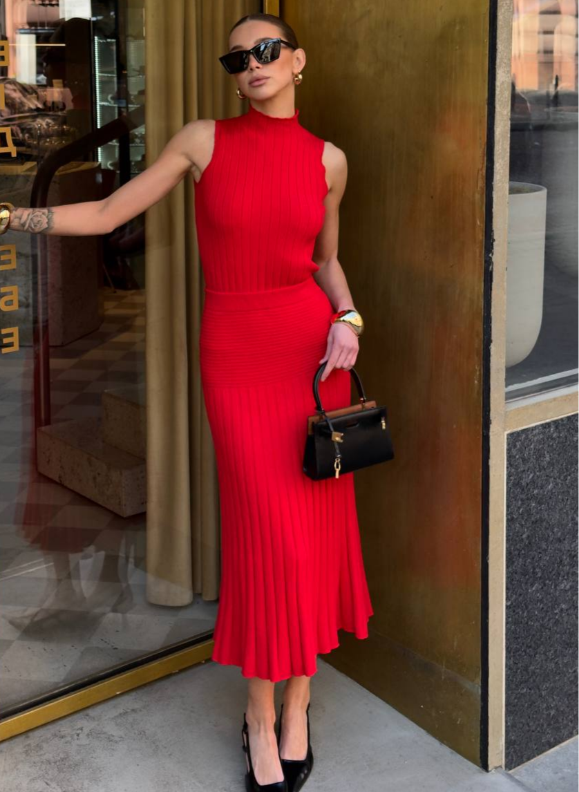Woman in a red dress holding a black handbag, standing in front of a reflective surface.